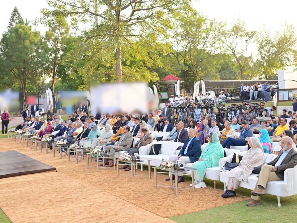 Guests seated on the lawn facing the stage, with a bank of TV cameras behind